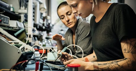 Female technician guiding young trainee in using a soldering iron.