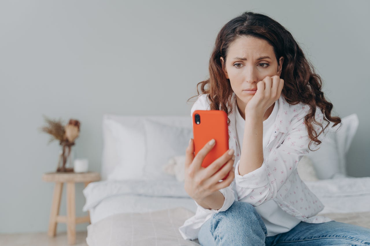 Young woman sitting on a bed, looking worried, staring at her smartphone