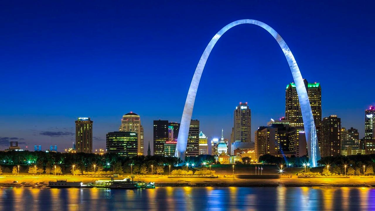 The St. Louis Gateway Arch at dusk, viewed from across the river. The sky is dark blue and the park around the Arch is lit with yellow light.