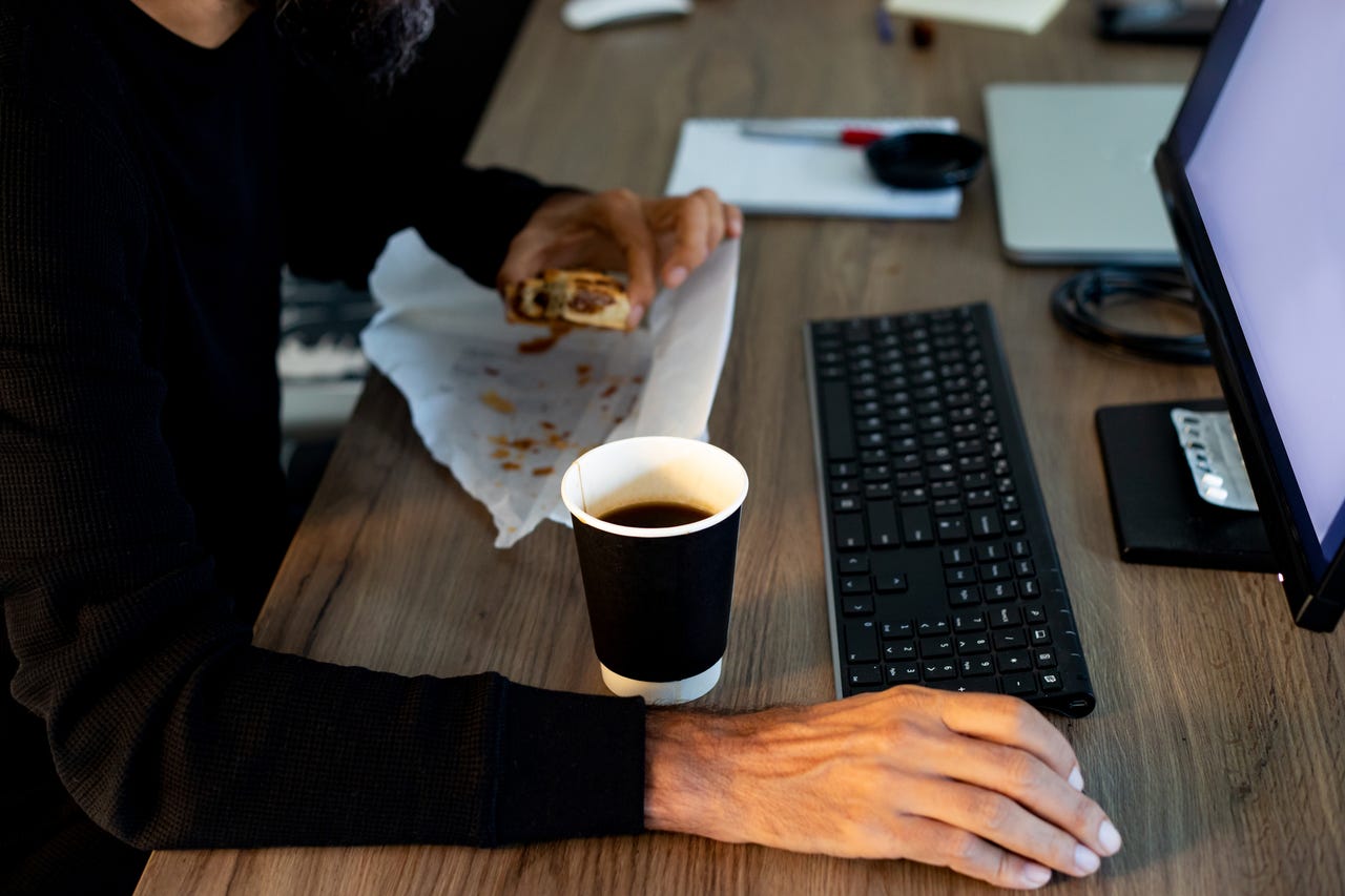 A person eating breakfast and drinking a coffee at their desk.