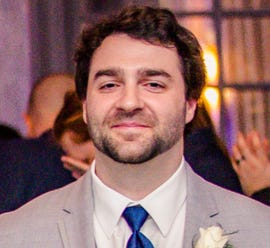 Foto kepala dan bahu seorang pria kulit putih berjanggut dalam setelan abu-abu dengan dasi biru.