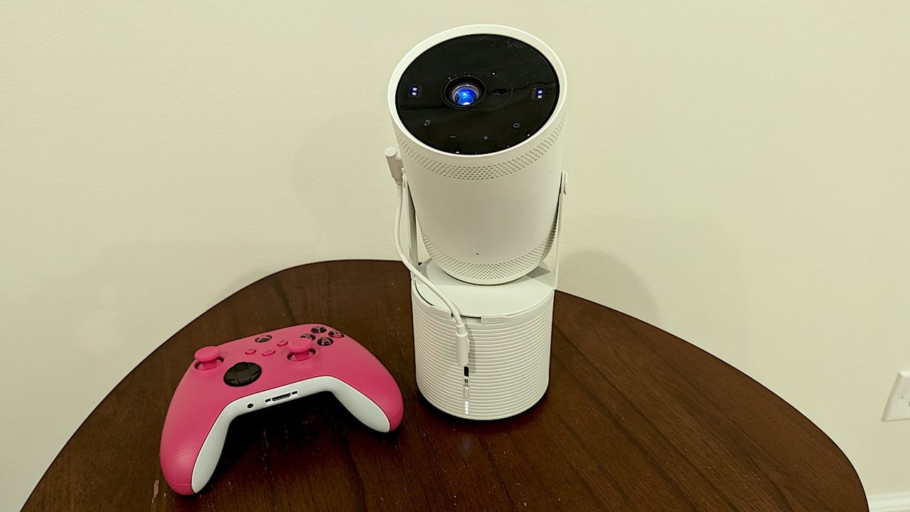 A white Samsung Freestyle 2 projector on its battery base next to a pink Xbox controller