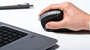Pessoa segurando um mouse preto ao lado de uma caneta e um laptop