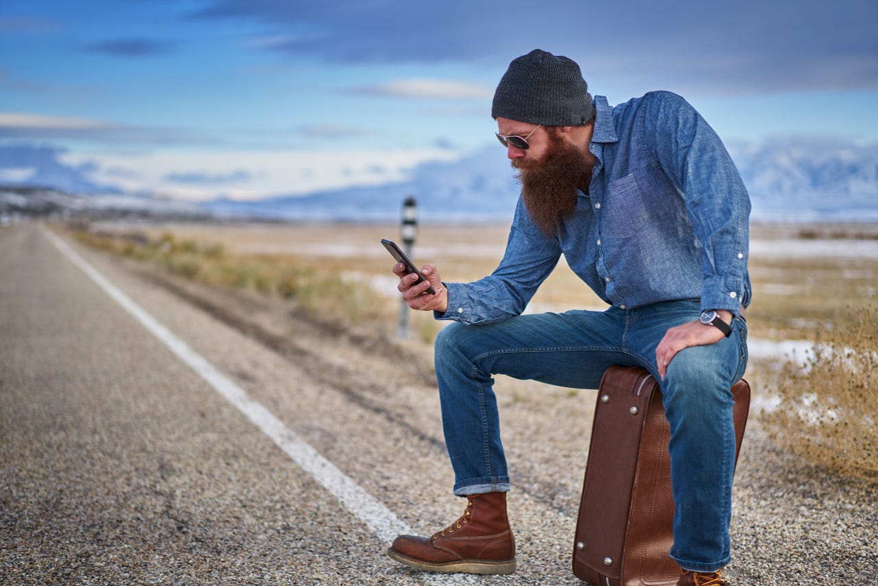 bearded hitch hiker sitting on suit case using smart phone