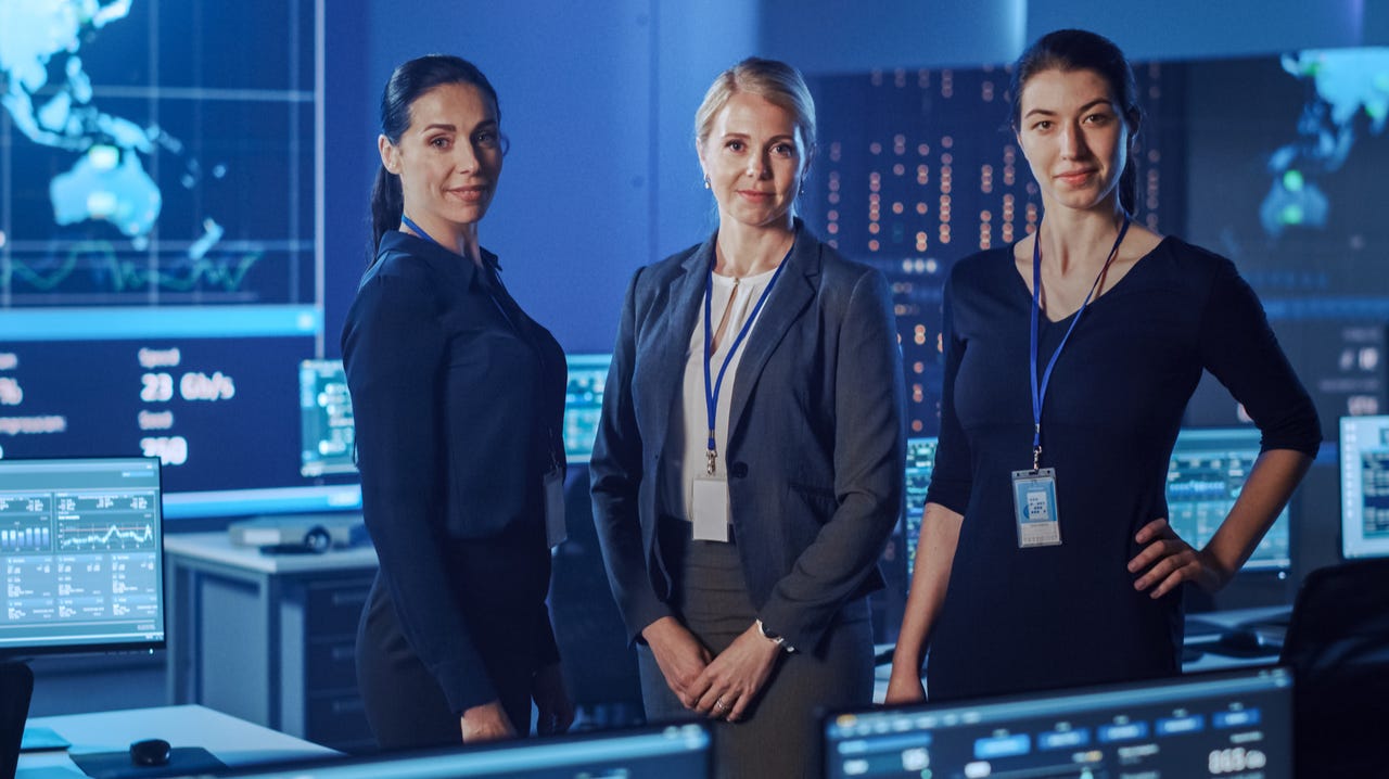 Three women wearing business clothes stand in a computer lab surrounded by screens.