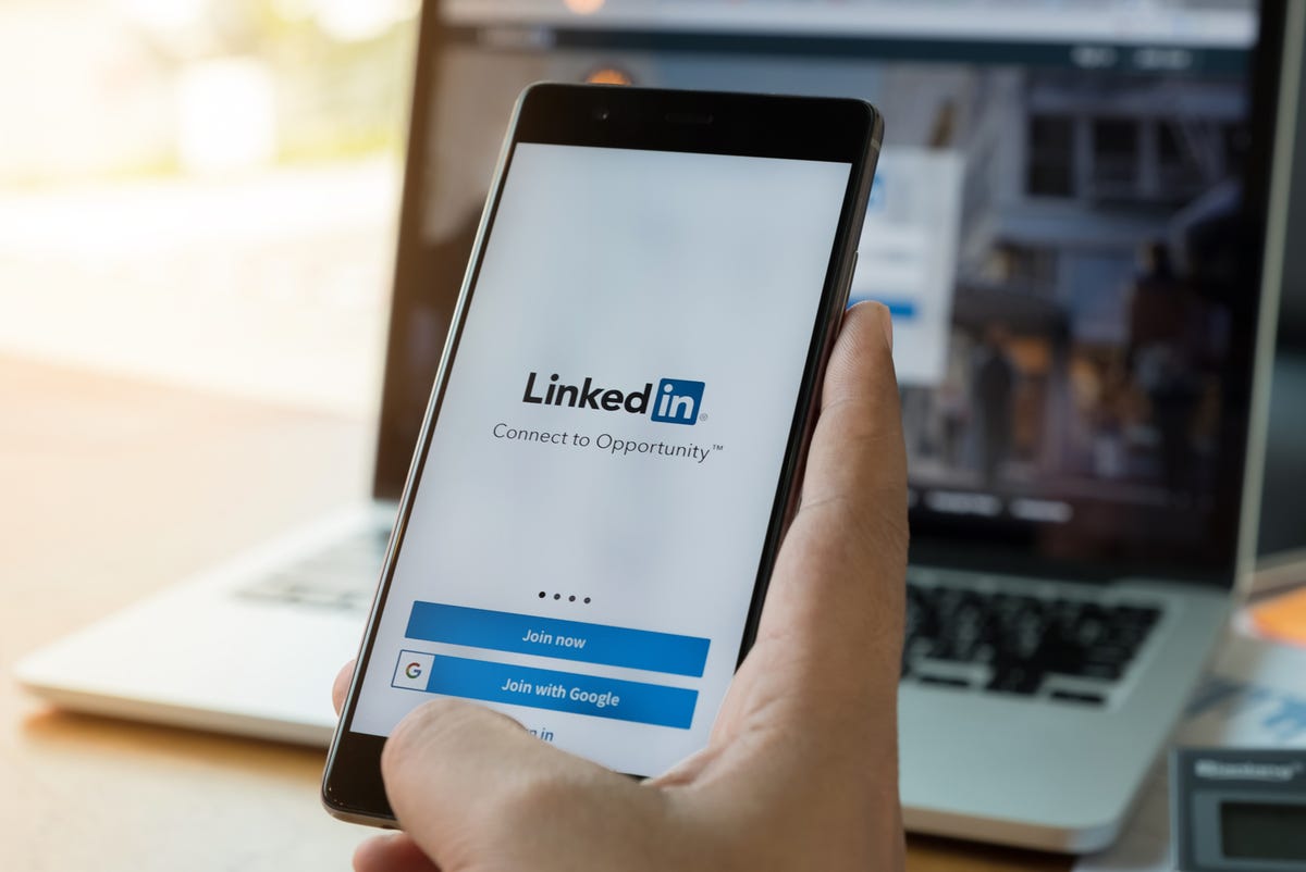 Hand holding a phone showing a LinkedIn login page, in front of an open laptop