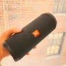 Hand holding JBL Flip 5 Portable Bluetooth Speaker against the backdrop of a brick building.