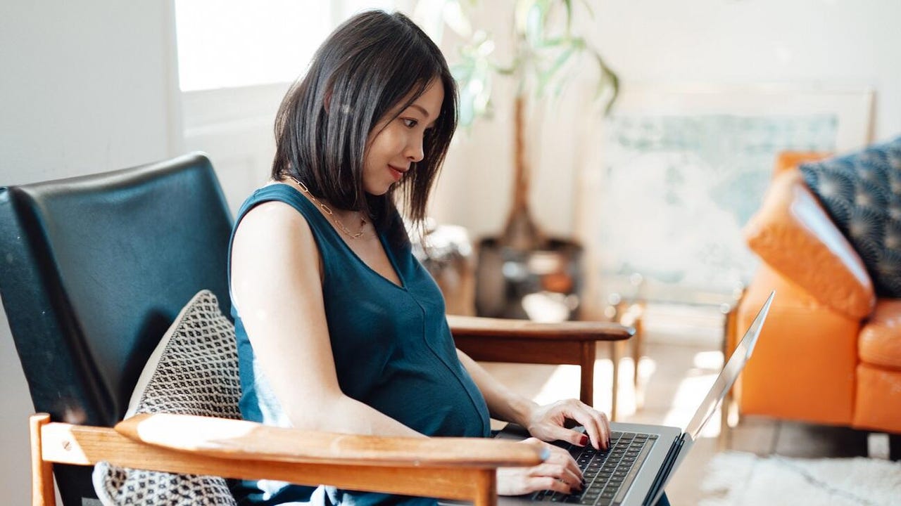 A pregnant Asian woman uses a laptop in a brightly lit, modern living room.