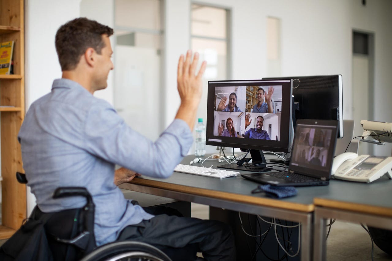 A middle aged man in a wheelchair sat at his computer desk, waving at smiling colleagues who are participating in a video call