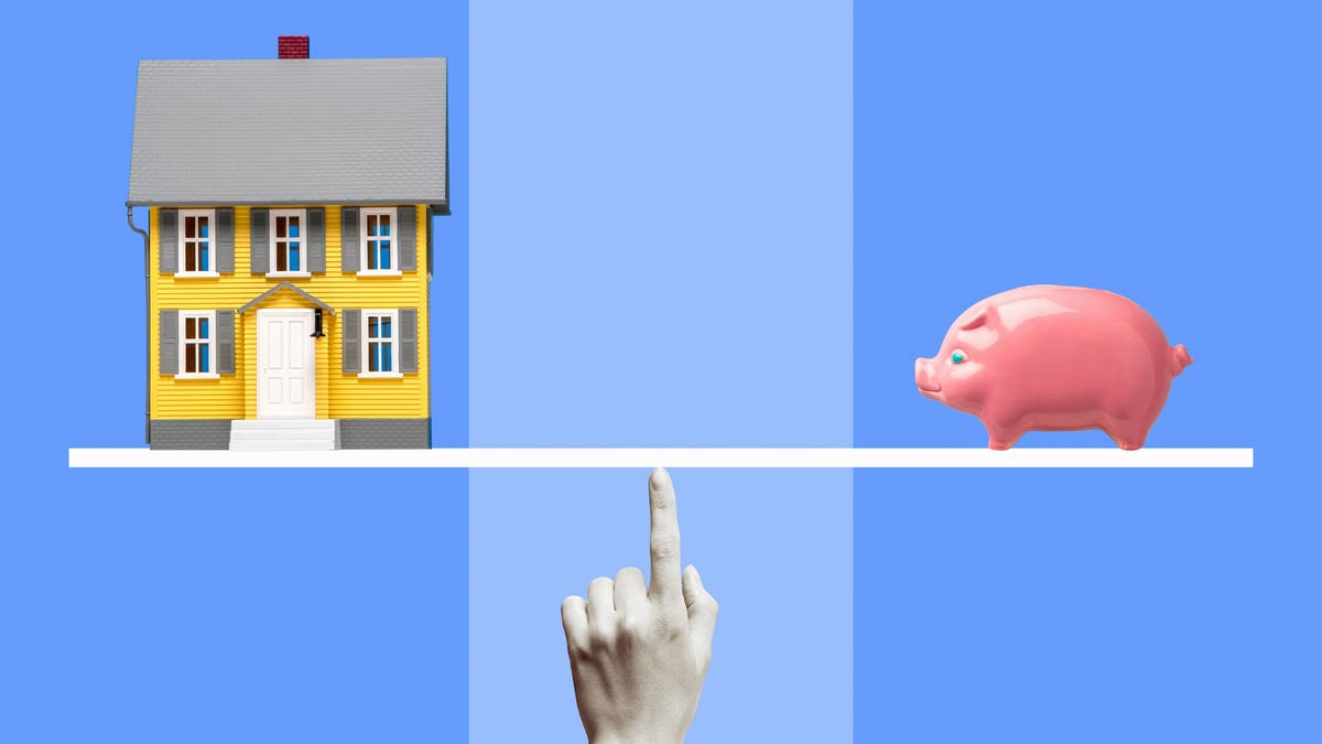 Yellow miniature model house and pink ceramic piggy bank on white line balanced on black and white woman's finger, blue background.
