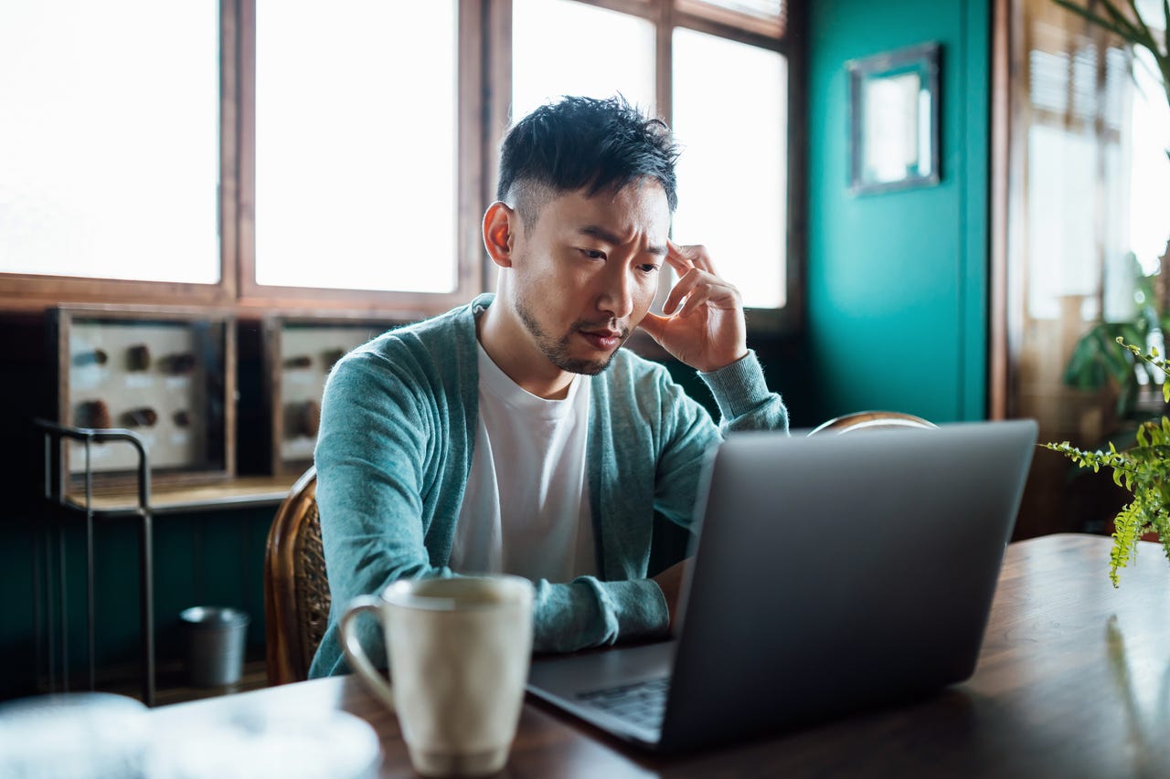 Worried young Asian man with his hand on head, using laptop computer at home