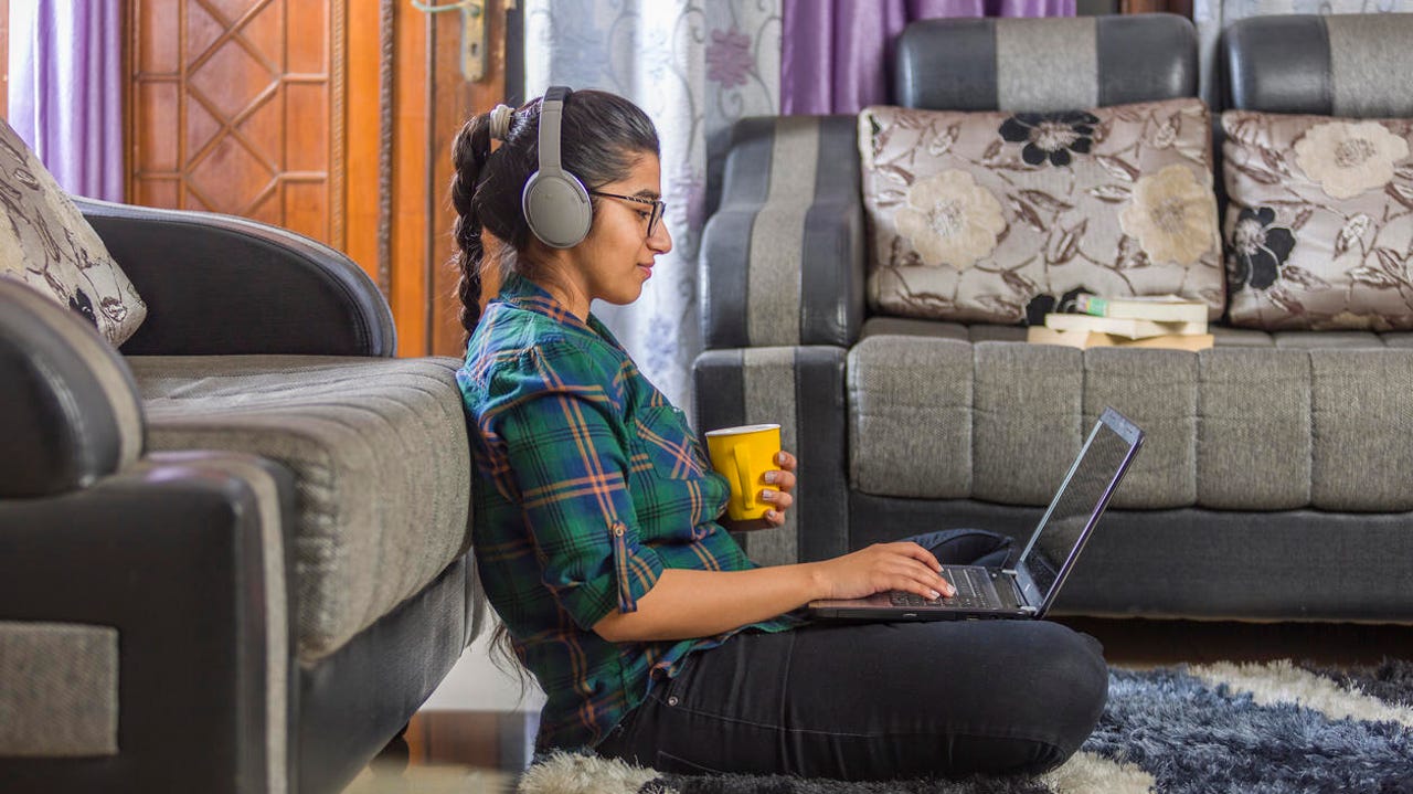 An Indian woman using laptop, sitting on the floor and leaning against a couch.
