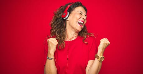 Middle age senior woman wearing headphones listening to music over red isolated background very happy and excited doing winner gesture with arms raised, smiling and screaming for success. Celebration concept.