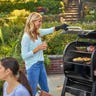 A man and woman stand next to a Weber SmokeFire EX4 pellet grill while another woman sits in the foreground, talking with a man off-screen.