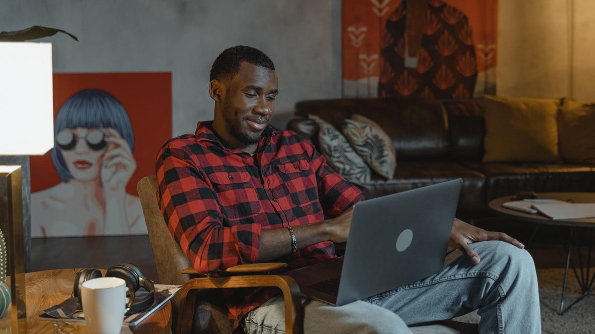 A Black man wearing a flannel shirt uses a laptop. He is sitting in a living room decorated with contemporary art.