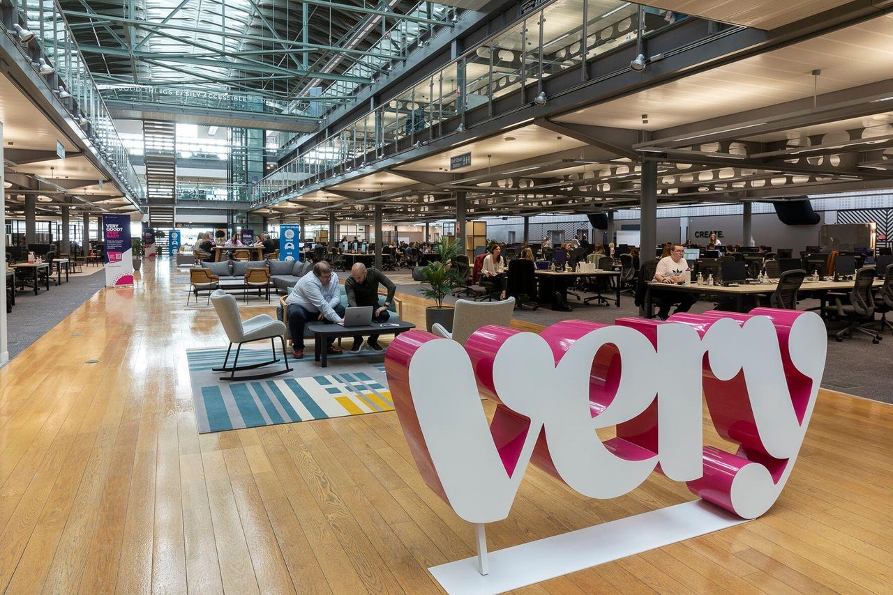 A white 'Very' logo set in the foreground of a spacious, modern-looking office
