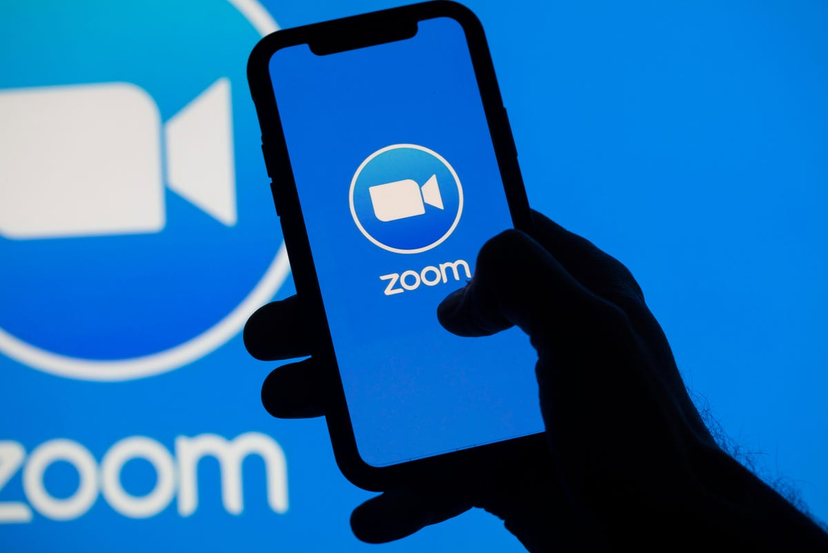 silhouette of a hand holding a mobile phone with the Zoom app displayed, in front of a blue background with the Zoom logo