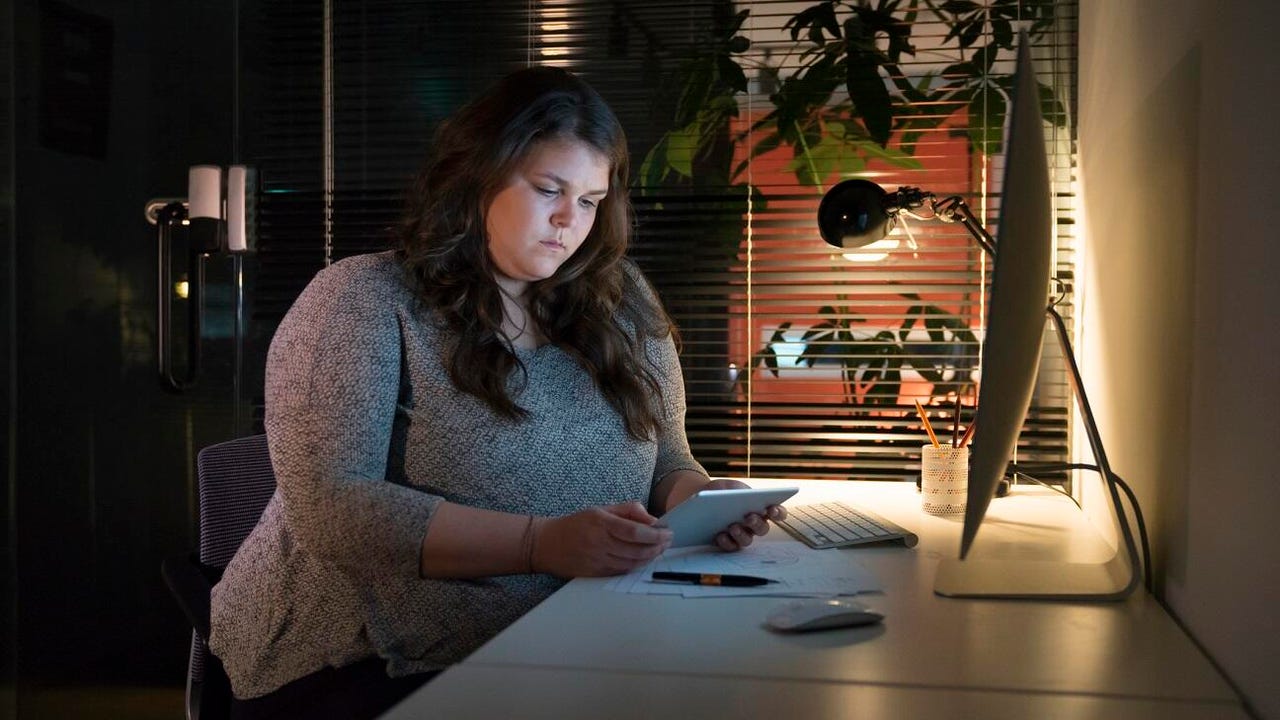 A female business professional works late. She is in a dark room sitting at a desk and looking at a tablet.