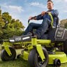 A man in work clothes using a Ryobi electric zero turn mower to cut his grass next to a flower bed