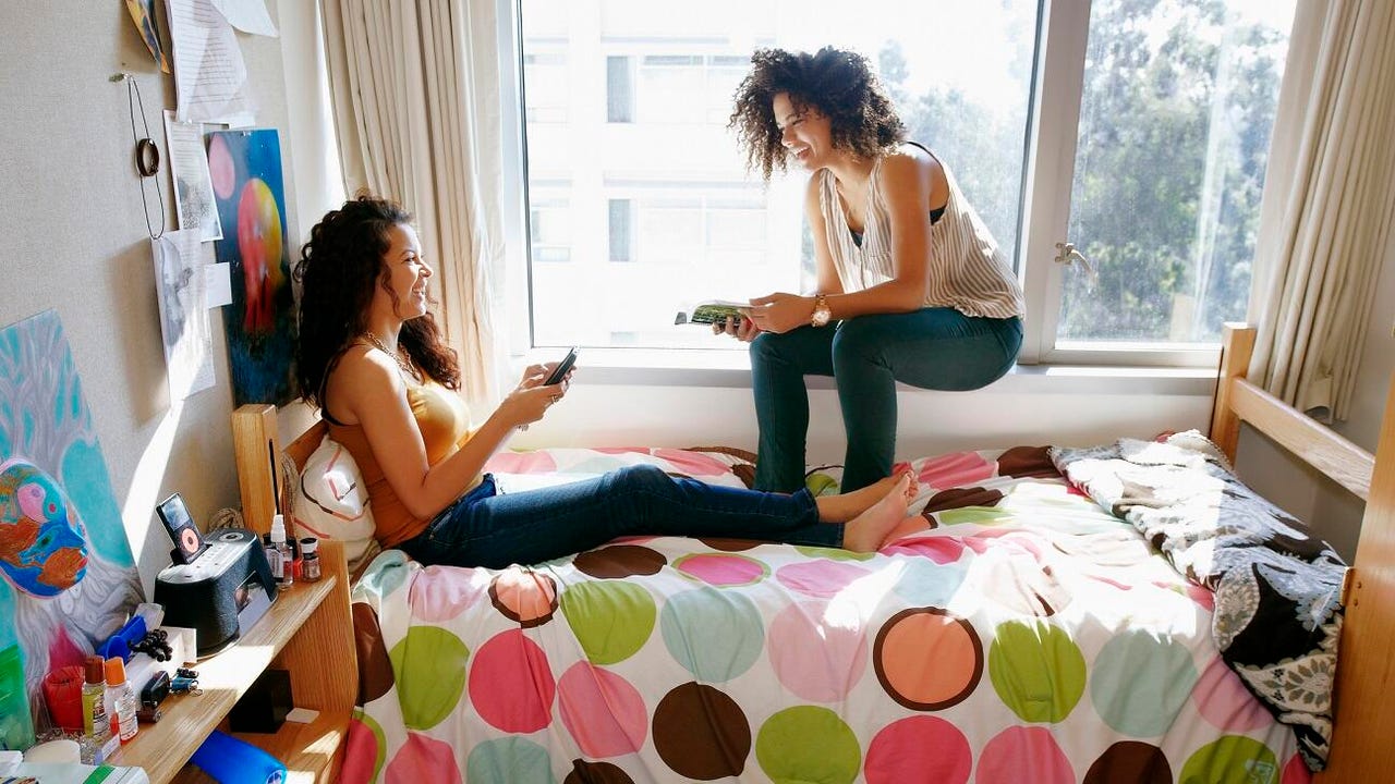 Two female college students with natural curly hair perch on a college dorm room bed, holding their phones. The room is colorful and cluttered.