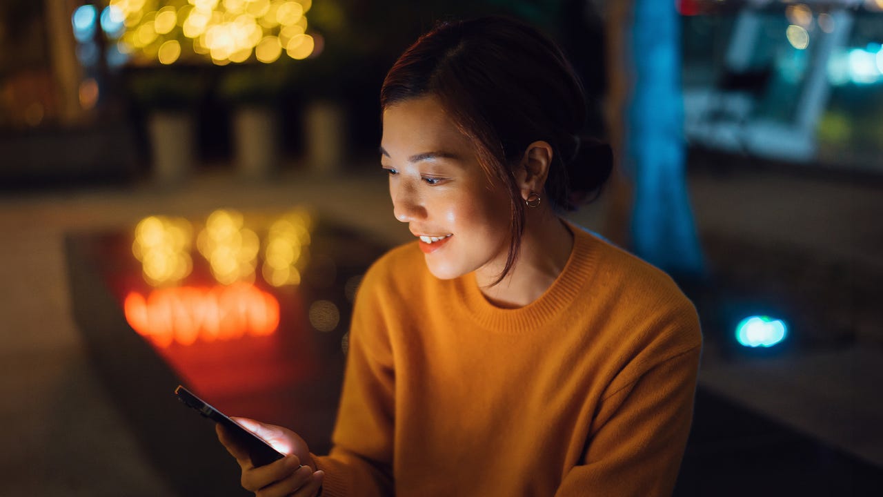 Beautiful smiling young Asian woman sitting in a pub and relaxing, surfing on the net and checking social media on smartphone. Spending a night out and enjoying city night life. Lifestyle and technology - stock photo