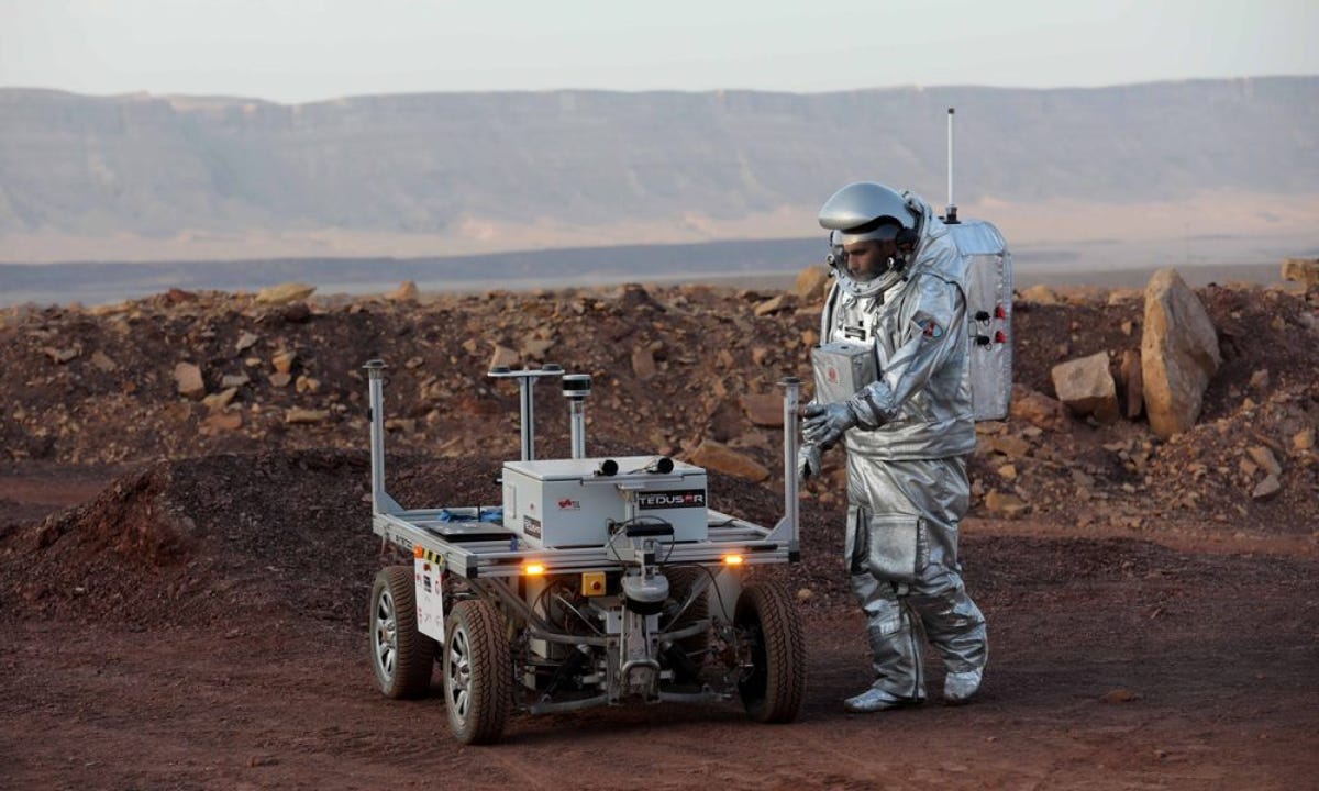 Astronaut and small wheeled vehicle in a desert setting with water in the background