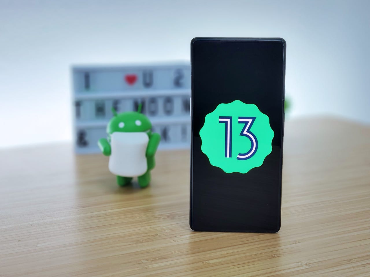 Android 13 logo on a phone on a table with small Android robot in the background.