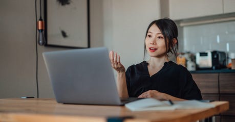 A confident Asian woman wearing business clothes speaks toward her laptop in her home office.