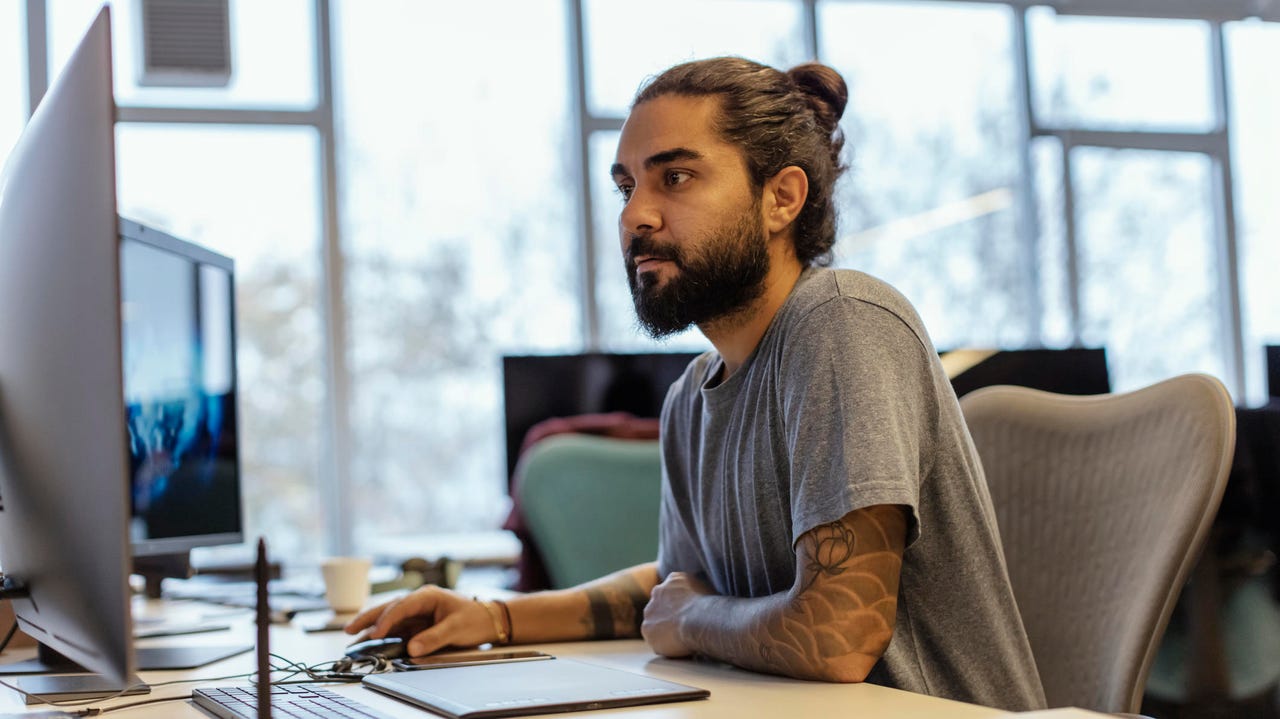 A software developer with tattoos, a beard and his hair in a bun sits at a desk in a well-lit office, looking at his dual computer screens.