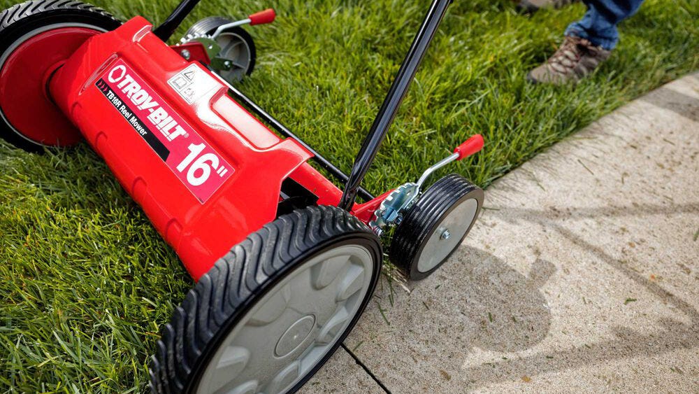 Partial view of someone using a Troy Bilt reel mower to cut grass next to a sidewalk.