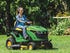 A woman sitting on a John Deere lawn mower, cutting grass near some flower beds on a sunny day