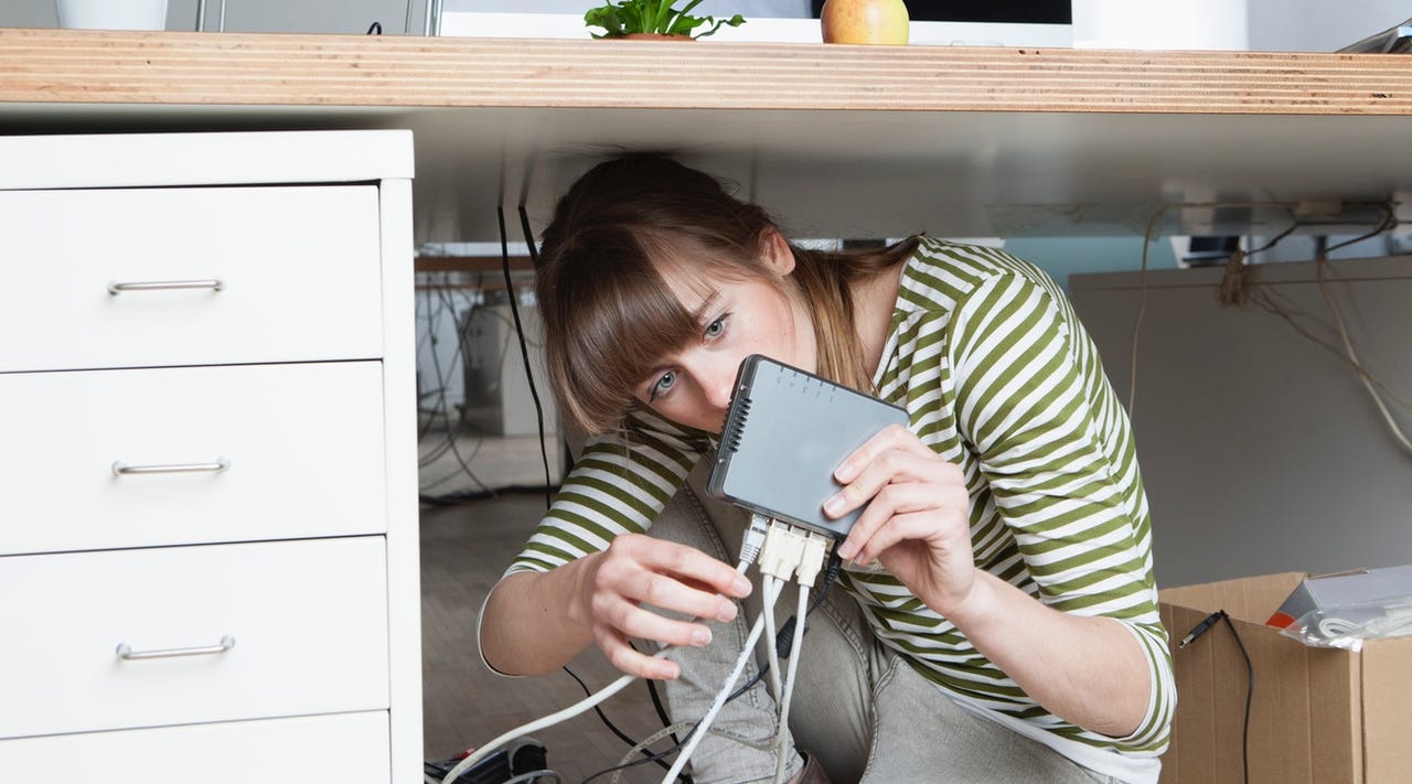 A woman crouched under a desk adjusting the many plugs in a router