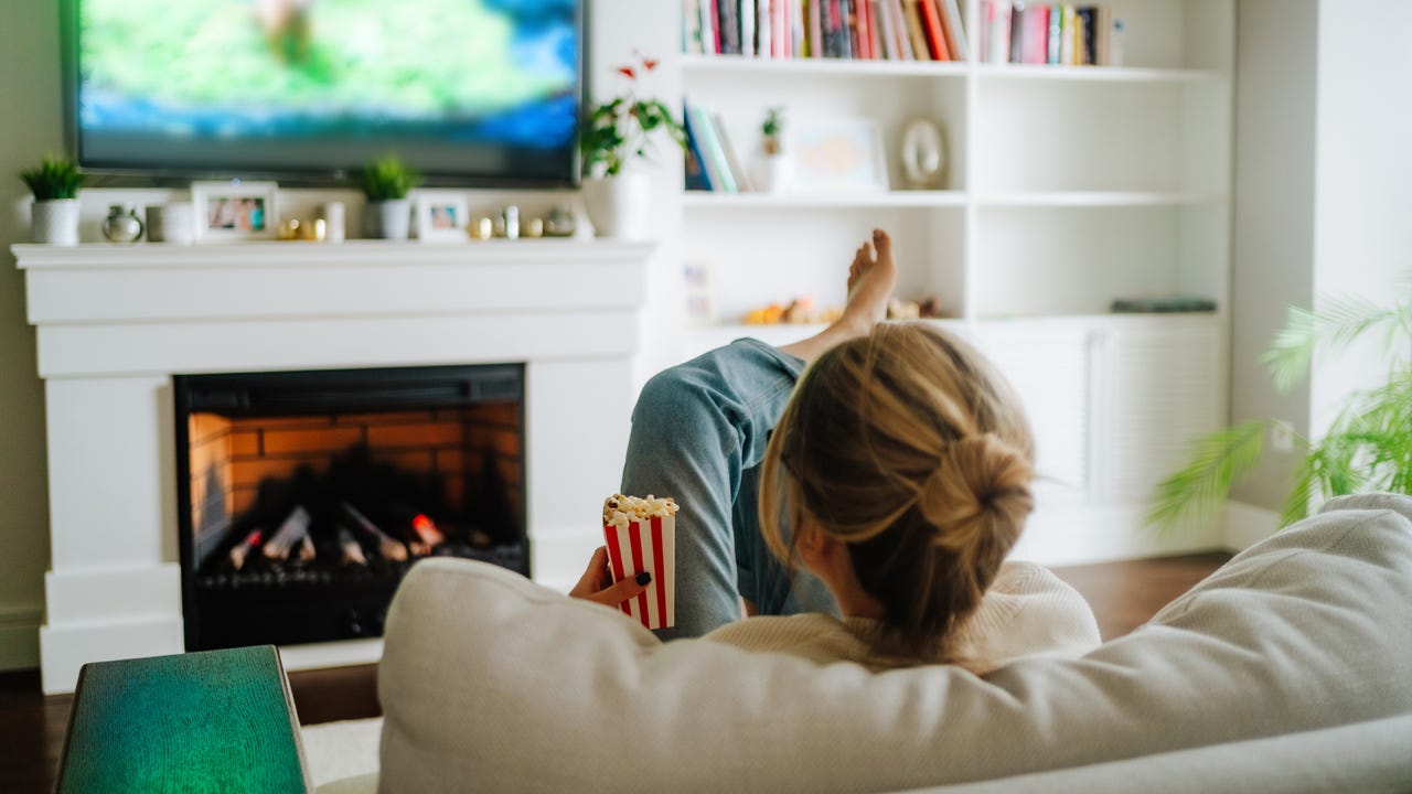Mujer rubia con moño viendo la televisión en un acogedor sofá frente a la chimenea en el interior