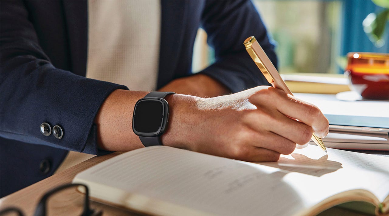 Close-up of a Fitbit Sense 2 smartwatch on the wrist of a young person writing in a journal