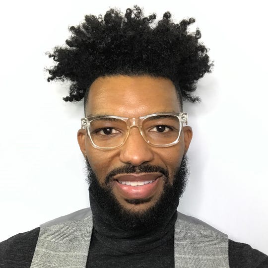 A head-and-shoulders photo of a Black man wearing large glasses, a black turtleneck, and a gray vest.
