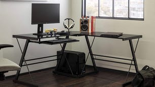 Black L-shaped desk with desktop monitor and speaker on it next to a window