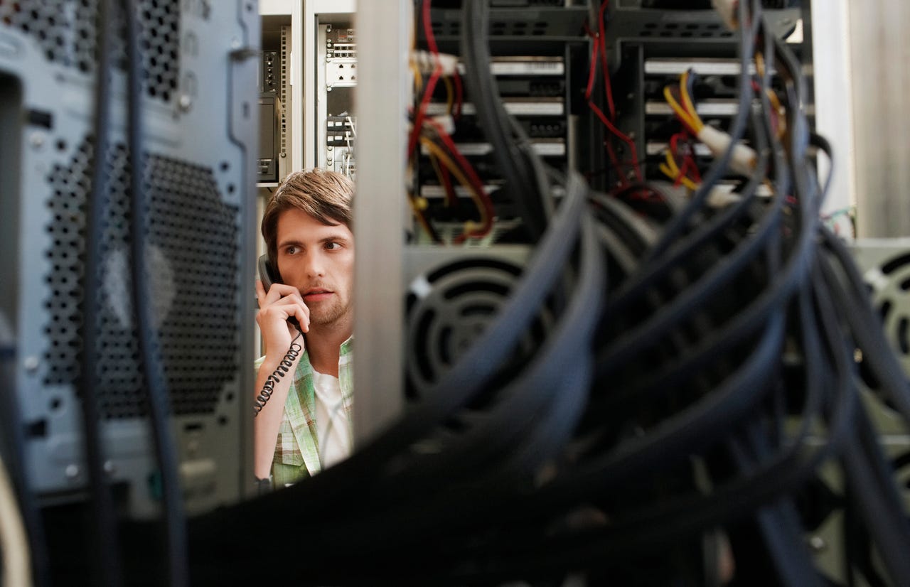 A male IT worker standing between racks of computers, speaking on the telephone with a serious expression