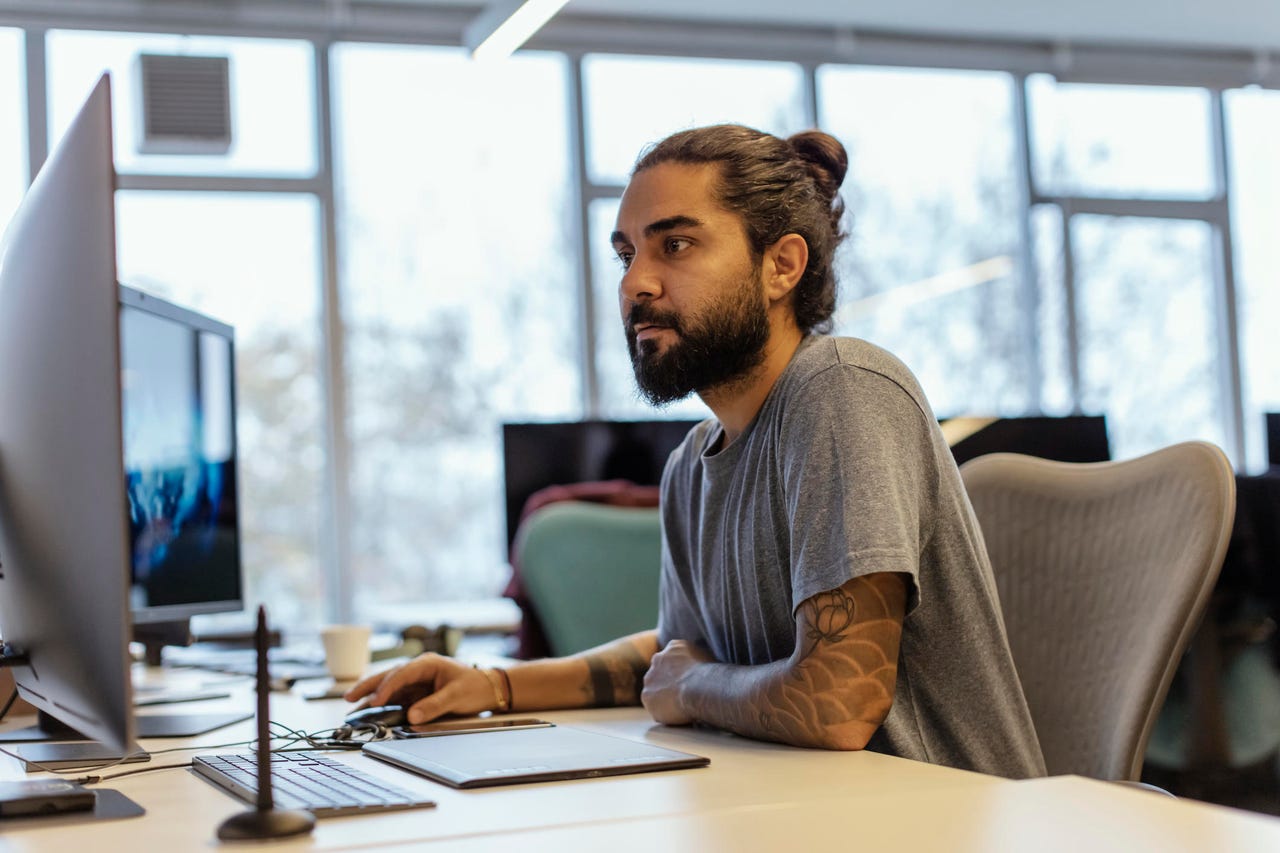 A software developer with a beard and his hair in a bun sits at a desk in a well-lit office, looking at his dual computer screens.