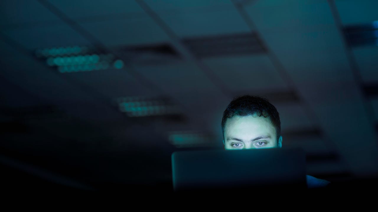 Eyes of a man  peering over the top of a laptop screen working late into the night with a dark empty office behind him