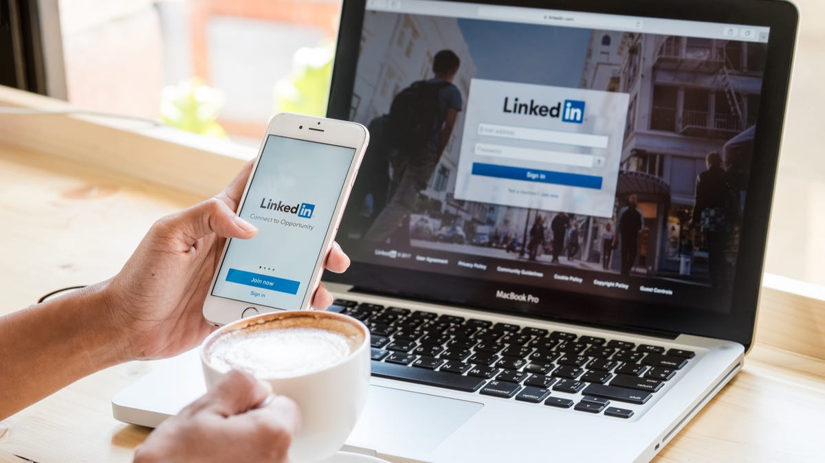 A hand holding a phone in front of a laptop, both showing LinkedIn's sign in page
