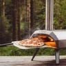Image of a person's hand removing a pizza from Ooni Karu 12 Multi-Fuel Pizza Oven on a wooden surface with a forest backgorund.
