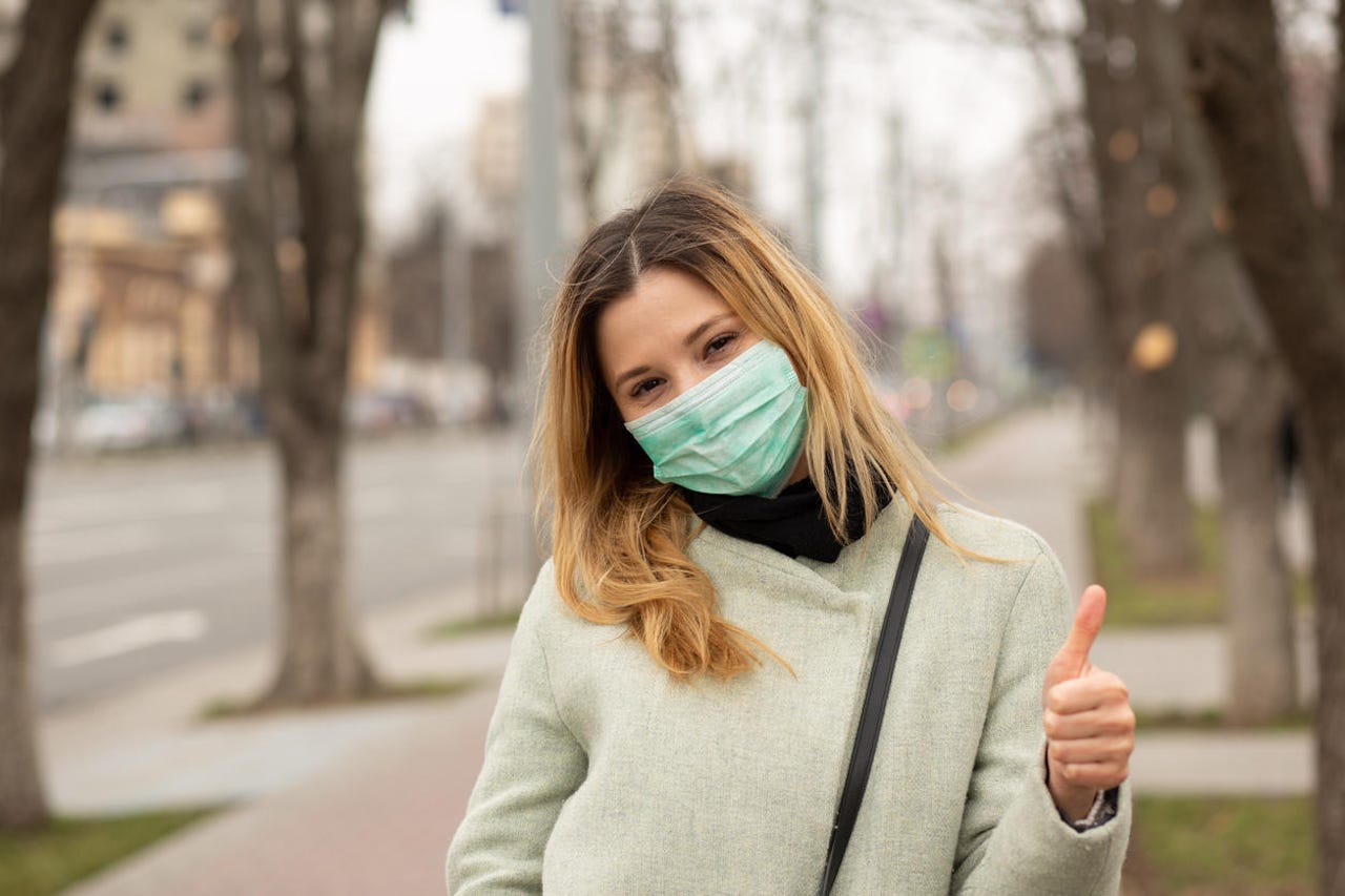 young woman in medical sterile protective mask on her face, walking in a European city, showing thumbs up