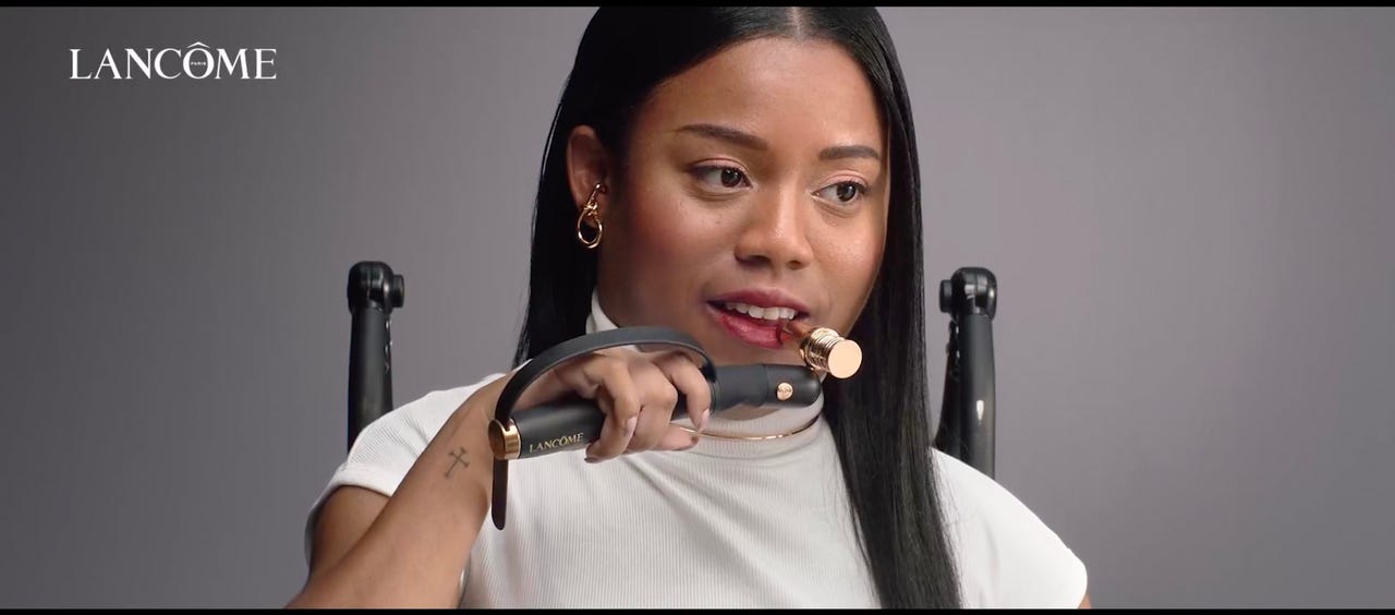 A young woman of color in a wheelchair with cerebral palsy uses the HAPTA lipstick applicator