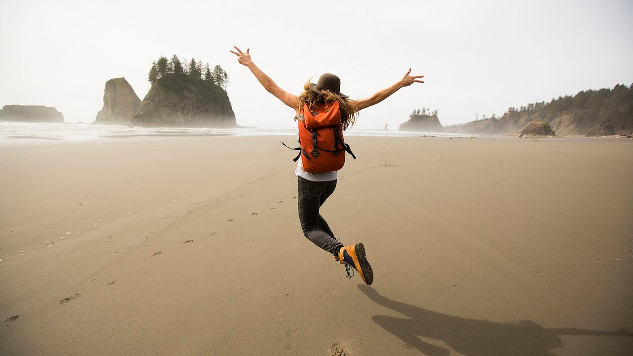 A long-haired person in hiking clothes leaps into the air on a beach in Washington.