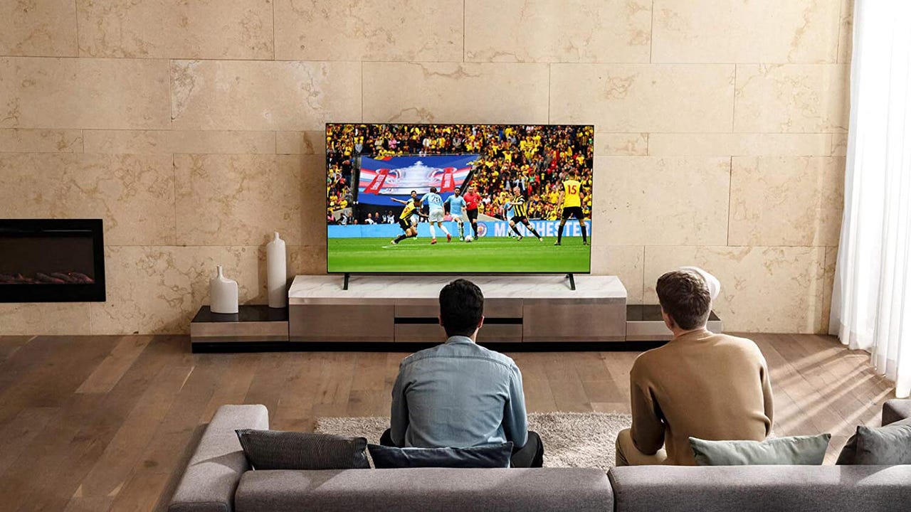 A from-behind view of two men sitting on a couch and watching a soccer match on an LG NanoCell 80 Series TV.