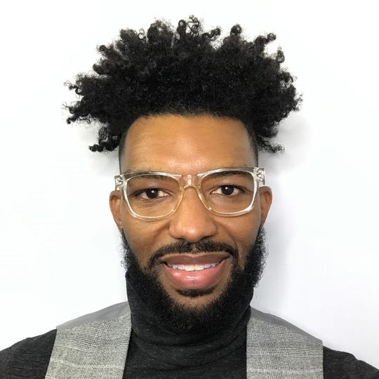 A head-and-shoulders photo of a Black man wearing large glasses, a black turtleneck, and a gray vest.