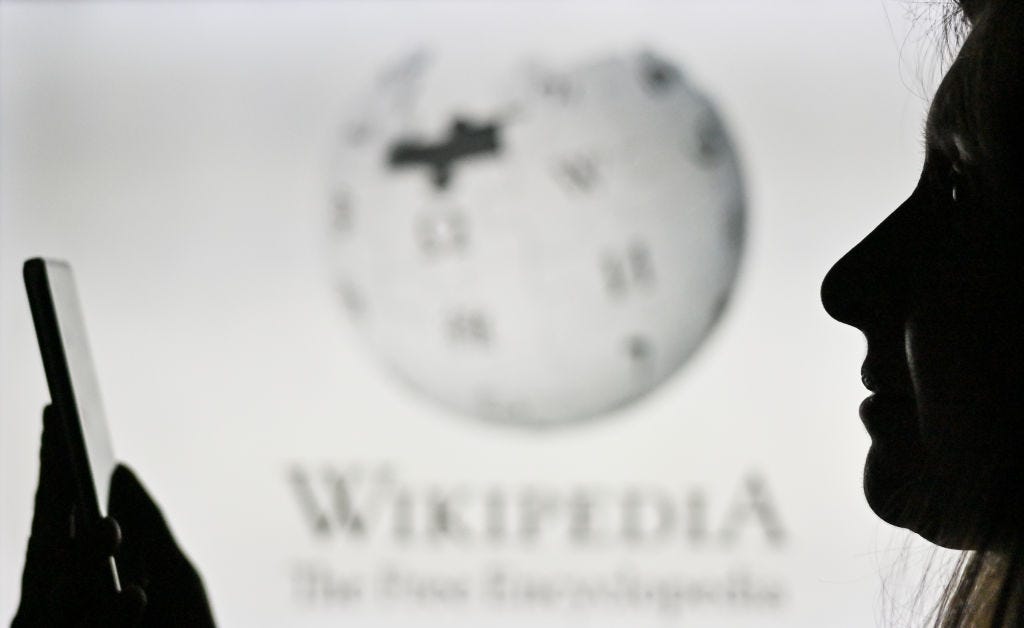 Image of a woman holding a mobile phone in front of the Wikipedia logo displayed on a computer screen.