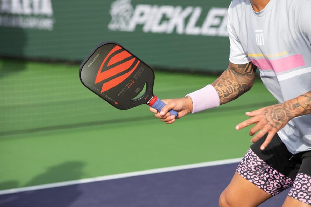 A pickleball player with paddle, colorful shorts, and sleeve tattoos in a sponsored arena