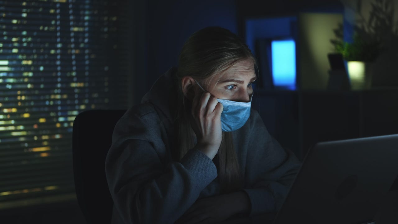 Close-up shot of Scared woman in medical mask reading news about coronavirus on laptop in darkness