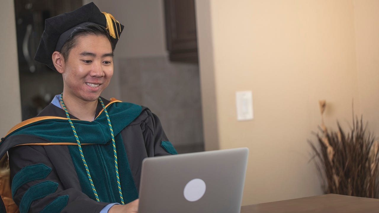 An Asian man celebrating his doctorate degree graduation while looking at a laptop.
